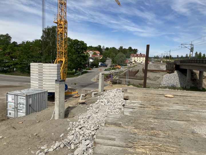 Vy över brobyggnationen vid Fridhemsgatan och Kyrkbergsvägen. Grundläggning för brostöd med armering syns intill befintlig järnvägsbro. Betongpålar i marken för förstärkning där marken för en arbetsyta förberetts. En byggkran, traktor och lastbil syns i arbetsområdet.
