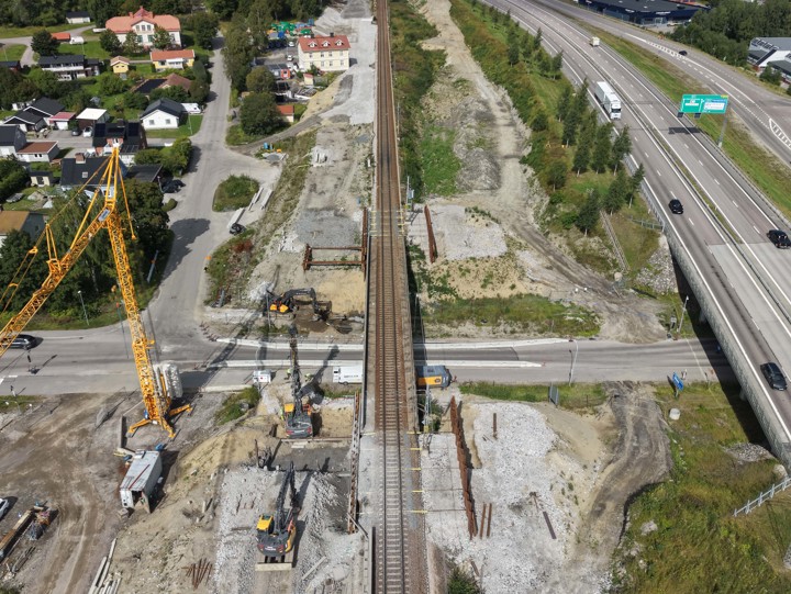Vy över brobyggnationen vid Fridhemsgatan och Kyrkbergsvägen. Grundläggning för brostöd med armering syns intill befintlig järnvägsbro. Betongpålar i marken för förstärkning. En byggkran och flera maskiner syns i arbetsområdet. På ena sidan järnvägsbron löper E4 och på den andra sidan ligger ett bostadsområde.