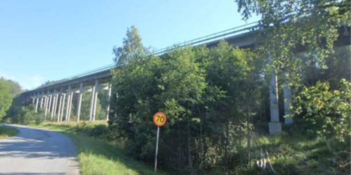 Delar av Gålåbron syns bakom grönskande träd. En väg passerar intill bron med en hastighetsskylt
