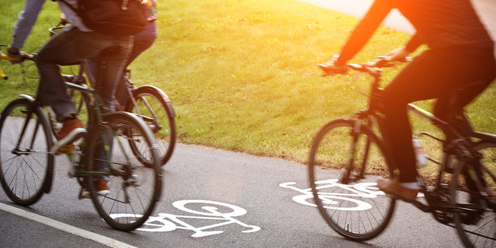 Tre cyklister på en cykelväg men en målad heldragen linje och två cyklar, en i varje färdriktning. Intill cykelvägen är det gräs som lyser upp av solen.