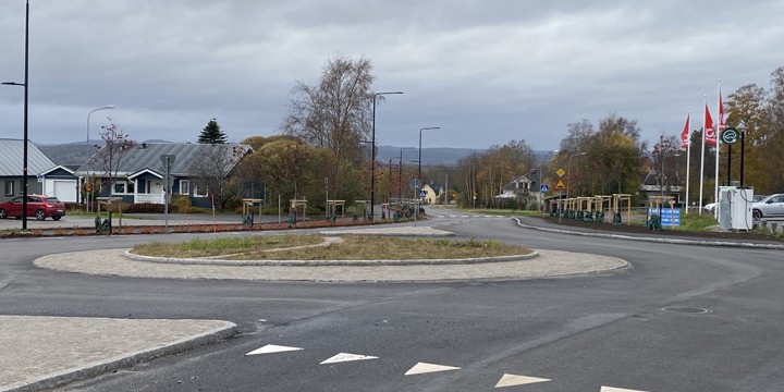 Centrumvägen i höjd med korsningen med Folketshusvägen med en färdig stenlagd cirkulationsplats. I mitten av cirkulationen växer gräs. vägen är asfalterad och målad. En privat fastighet syns till vänster och flaggor för en dagligvaruhandel till höger. Himlen är täckt av moln och träden skiftar i höstens färger.