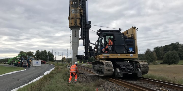Maskin som är placerad på järnvägsspår trycker ner en påle i marken bredvid spåret. Bilväg går till vänster.
