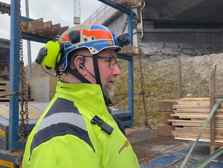 Man med grått skägg och glasögon står utomhus vid en väg med en lastbil i bakgrunden. Halvfigur men hjälm och gula arbetskläder.